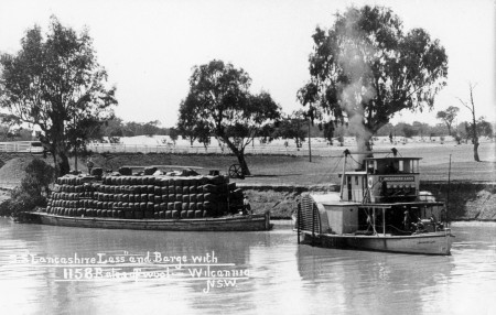 Black and white photograph of a paddle steamer travelling along a river pulling a heavily loaded barge